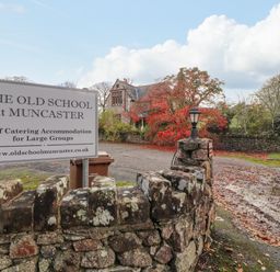 A stone wall with a sign for The Old School at Muncaster and a driveway leading to a building with trees in Ravenglass near Holmrook