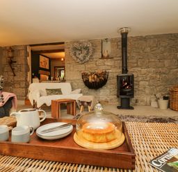 A living room with a stone wall and wood-burning stove with a tray of tea and cake on a wicker table at Lower Gadles Burnthouse near Falmouth