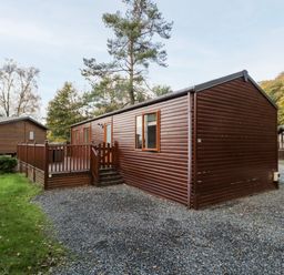 A brown wooden cabin with a fenced deck and gravel driveway surrounded by trees at Wansfell Lodge Pony Meadow 36 White Cross Bay near Windermere