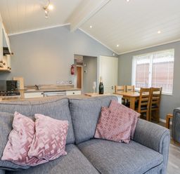An open living room and kitchen area with gray sofas with pink cushions a wooden dining table with chairs and kitchen appliances at Wansfell Lodge Pony Meadow 36 White Cross Bay near Windermere