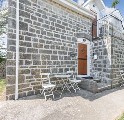 Exterior of a stone building with two white chairs and a table on each side of a wooden door at The Coach House in St Florence