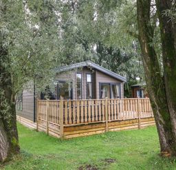 A wooden cabin with a fenced porch surrounded by trees and grass at Coniston View 9 Dock Acres near Tewitfield