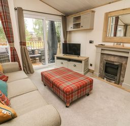A living room with a sofa with cushions a plaid ottoman a fireplace a television on a cabinet and sliding glass doors at Coniston View 9 Dock Acres near Tewitfield