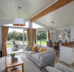 A living room with a grey sofa and yellow cushions next to sliding glass doors leading to a patio with outdoor seating at Fairview Lodge in Pwllheli