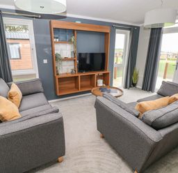 A living room with two grey sofas with yellow cushions a wall-mounted wooden shelving unit with a TV a round wooden coffee table and large windows at Driftwood Lodge in Hasguard Cross near Broad Haven