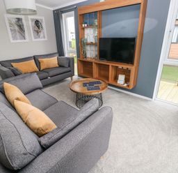A living room with grey sofas and brown cushions a round wooden coffee table and a wall-mounted wooden entertainment unit with a TV at Driftwood Lodge in Hasguard Cross near Broad Haven