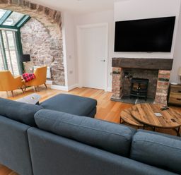 A living room with a blue sofa a round wooden coffee table a brick fireplace with a black stove two yellow chairs near a stone wall window at Skomer Cottage Sandy Haven near Milford Haven