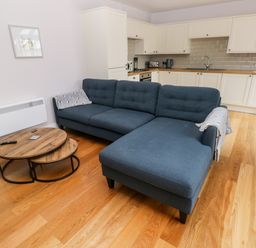 A living room with a blue sectional sofa a wooden nesting coffee table and a kitchen with white cabinets at Skomer Cottage in Sandy Haven near Milford Haven