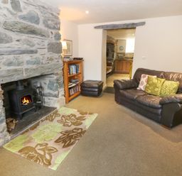 A living room with a stone fireplace and wood stove a leather sofa with cushions a wooden bookshelf and an open doorway leading to a kitchen at Tyn Llwyn in Eisingrug near Harlech
