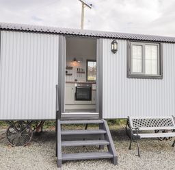 An exterior view of a small corrugated metal hut on wheels with steps leading to an open door and a bench nearby at Shepherds Hut in Dunvegan