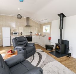 A living room with gray recliner chairs a kitchen area with white cabinets and appliances and a black wood stove at Cliff Barn in Burton Bradstock