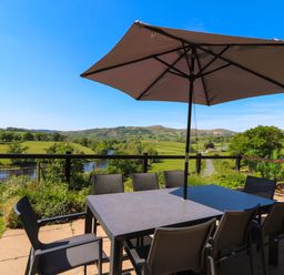 An outdoor patio table with chairs and an umbrella overlooking a river and green hills at Pen Y Banc in Llanfaredd near Builth Wells