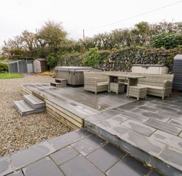 An outdoor patio area with wicker furniture table chairs hot tub storage sheds and gravel ground at Cae Du in Dwyran near Niwbwrch Newborough