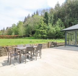An outdoor patio with a table and six chairs next to a stone wall and a building with glass sliding doors surrounded by trees at Coffey's Ballymacarbry County Waterford