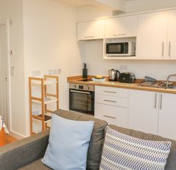A compact kitchen with white cabinets microwave oven toaster kettle and a sink next to a gray sofa with blue and patterned cushions at The Mew in Dartmouth