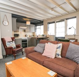 A living area with a brown sofa with cushions a wooden table and a leather armchair next to a kitchen at Paddock Cottage Nr Malmesbury
