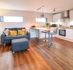 An open plan living room and kitchen with a grey sofa and ottoman a wooden floor and a kitchen island with bar stools at Meols Holiday Lodge in Meols
