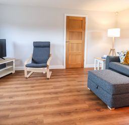 A living room with a gray armchair, a television on a white stand, a gray sofa with cushions, a gray ottoman, a wooden door, and a floor lamp at Meols Holiday Lodge in Meols