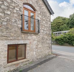 A stone exterior wall with two wooden framed windows next to a roadside with trees and bushes in the background at Coach House in Arnside