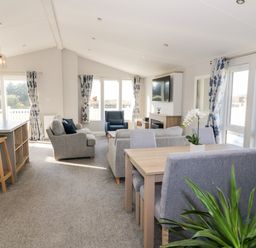 A living room and dining area with grey chairs a wooden table a kitchen island with stools and floral curtains at Dershel Lodge in Southerness