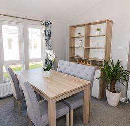 A dining area with a wooden table grey upholstered chairs a wooden shelving unit and a potted plant at Dershel Lodge in Southerness