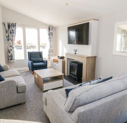 A living room with sofas a chair a coffee table a fireplace a television and windows with curtains at Dershel Lodge in Southerness