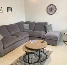 A living room with a grey sectional sofa a round wooden coffee table a small side table and a wall clock at LunaRoxx 6 Meadow Gardens Kilgetty