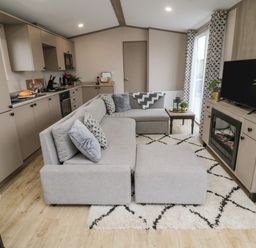 An open-plan living room and kitchen with a grey sectional sofa a patterned rug and a TV on a wooden cabinet at Plot 18 Meadow View in Felton