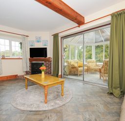 A living room with a wooden coffee table and a TV on a brick fireplace with a view into a conservatory with wicker furniture at The Wagon House in Lezant near Launceston