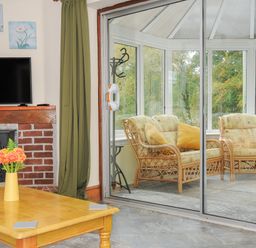 A living room with a coffee table and fireplace adjacent to a conservatory with wicker chairs and large windows at The Wagon House in Lezant near Launceston
