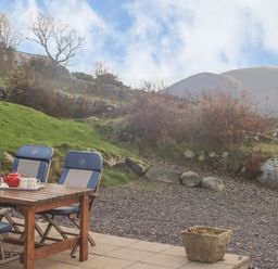 An outdoor patio with a wooden table and cushioned chairs with a teapot and cups on the table and a hillside with rocks and bushes in the background at Beech Tree Cottage in Waunfawr