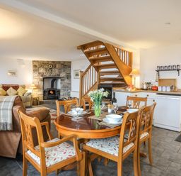 A dining area with a wooden table set with dishes and glasses next to a kitchen and a living room with a stone fireplace and wooden staircase at Beech Tree Cottage in Waunfawr