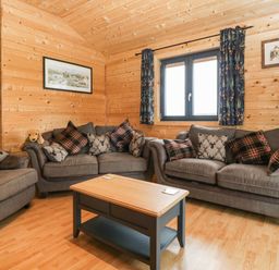 A living room with wooden walls two gray sofas patterned cushions a wooden coffee table and a window with curtains at The Lodge at Crossroads Farm Middlezoy