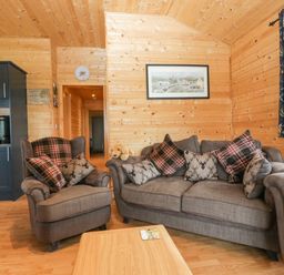 A living room with grey armchairs and sofa with patterned cushions wooden walls and flooring a blue kitchen unit and a wooden coffee table at The Lodge at Crossroads Farm in Middlezoy