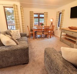 A living room with grey sofas, wooden dining table and chairs, a sideboard, and a wall mounted TV at Woodland Retreat Skiptory Howe 41A White Cross Bay near Windermere