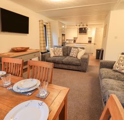 A living room with a dining table set with plates and glasses two sofas with cushions a wall-mounted TV and a view of the kitchen at Woodland Retreat Skiptory Howe 41A White Cross Bay near Windermere