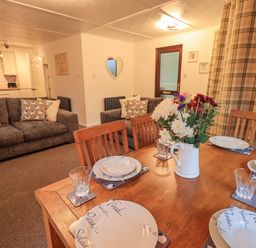 A dining table set with plates glasses and a vase of flowers next to a living area with sofas and cushions at Woodland Retreat Skiptory Howe 41A White Cross Bay near Windermere