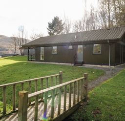 A wooden cabin with a tiled roof near a small wooden bridge over a grassy area at Firbush Lodge in Killin