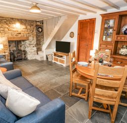 A living room with a blue sofa a stone fireplace a wood burning stove a wooden dining table and chairs and wooden cabinets at Dunnock Cottage in Broad Haven
