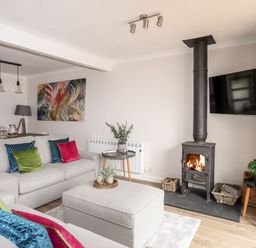 A living room with a grey sofa and cushions a wood burning stove a wall-mounted TV and a dining table at 240 Cae Du in Abersoch