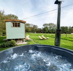 An outdoor bubbling hot tub with a wood chimney and a small green cabin and two wooden lounge chairs on grass at Fern in Broad Langdon near Wainhouse Corner