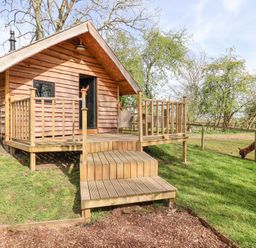 A cabin with a small wooden deck and steps surrounded by grass and trees with an animal grazing at Duck Lodge in Bedale
