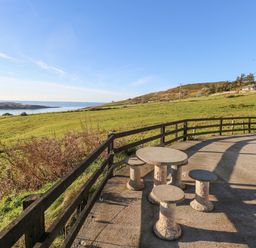 A patio area with a round stone table and stools overlooking green fields and water at Castle Demesne House in Clifden County Galway
