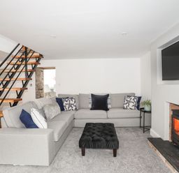 A living room with a grey sectional sofa and black ottoman near a fireplace and a staircase at Bodwena in Moelfre