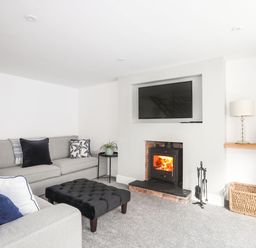 A living room with a grey sofa a black ottoman a fireplace with a wood-burning stove a television a lamp books and a wicker basket at Bodwena in Moelfre