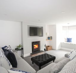 A living room with a wood burning stove a wall mounted tv a grey sofa a black ottoman and a window seat at Bodwena in Moelfre