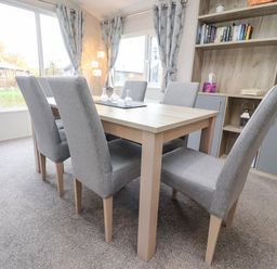 A dining area with six grey upholstered chairs around a wooden table near windows and a shelving unit at Fountain View in Warton near Carnforth