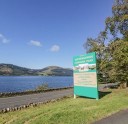 A road next to a body of water with a green sign for Auchengower Caravan Park and trees at 11 Mansion View in Helensburgh near Kilcreggan