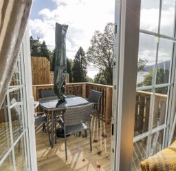 A balcony with a table and four chairs surrounded by wooden railings and trees visible through open glass doors at 11 Mansion View in Helensburgh near Kilcreggan