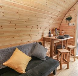 A wooden interior with a black couch with cushions and a small dining table with stools beside a door open to the outside at Parys Pod in Amlwch